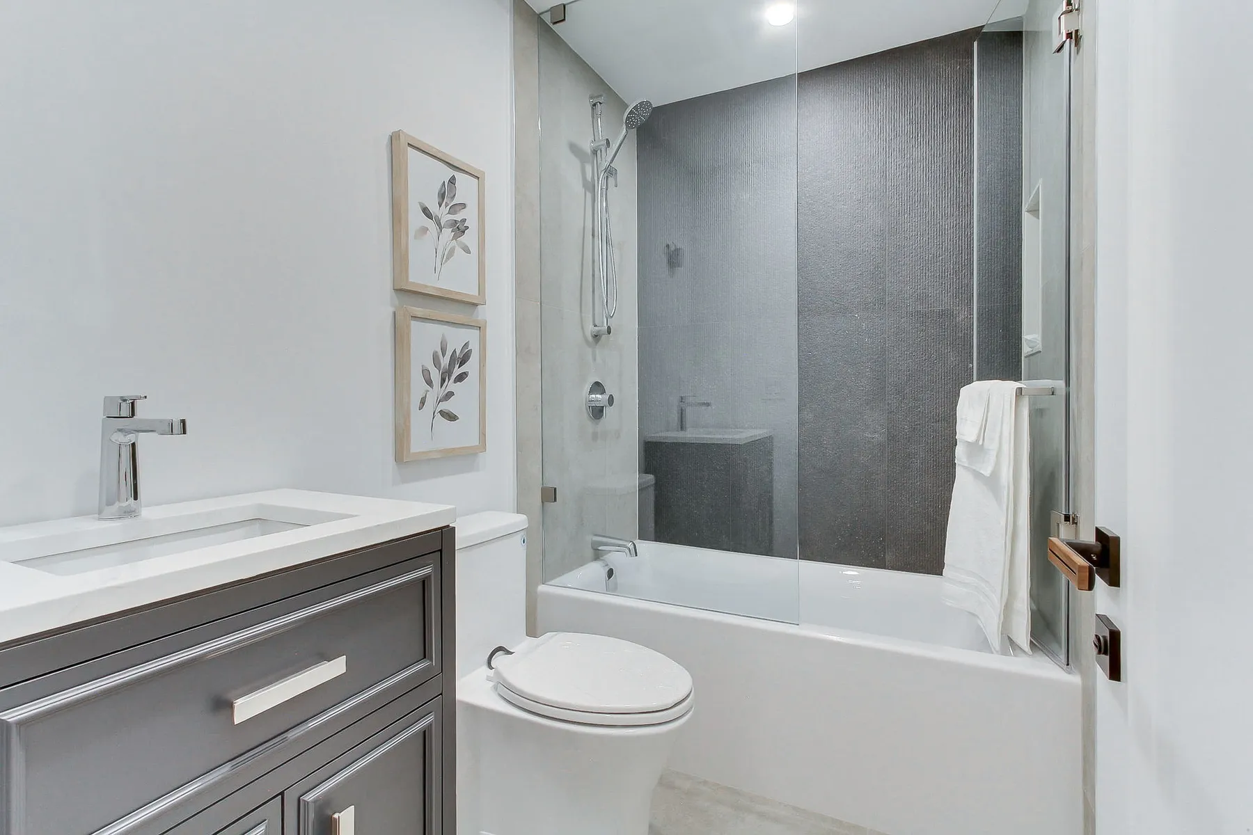A full bathroom with a grey vanity, quartz top and a tub-shower clad in dark large-format tile