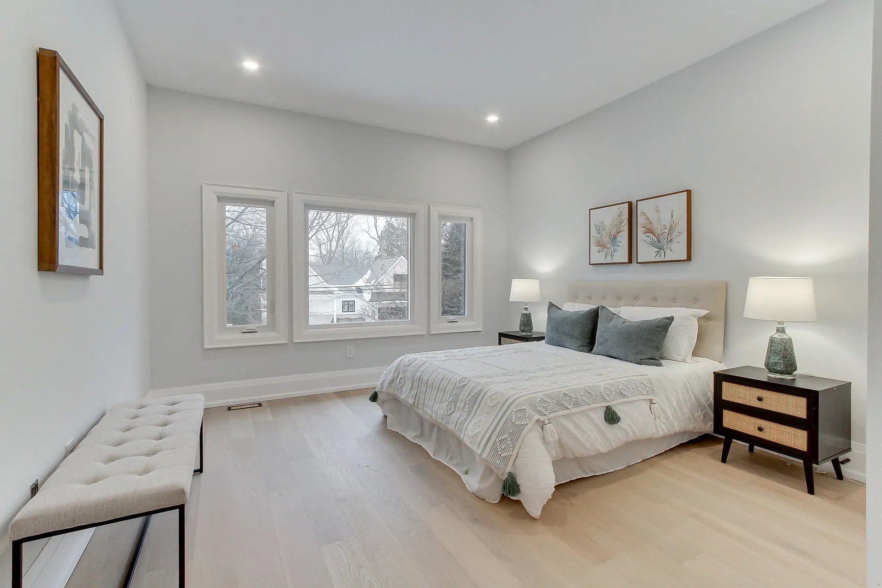 A primary bedroom with a tufted cream headboard, a wide triple window and light oak floors