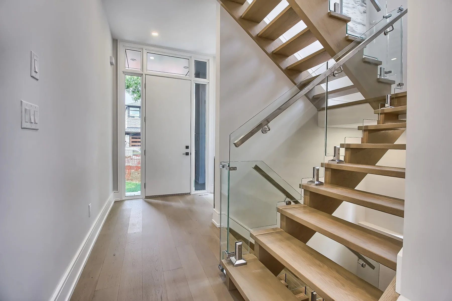 The entry hall with a white-oak open-riser staircase and a glass railing beside the front door