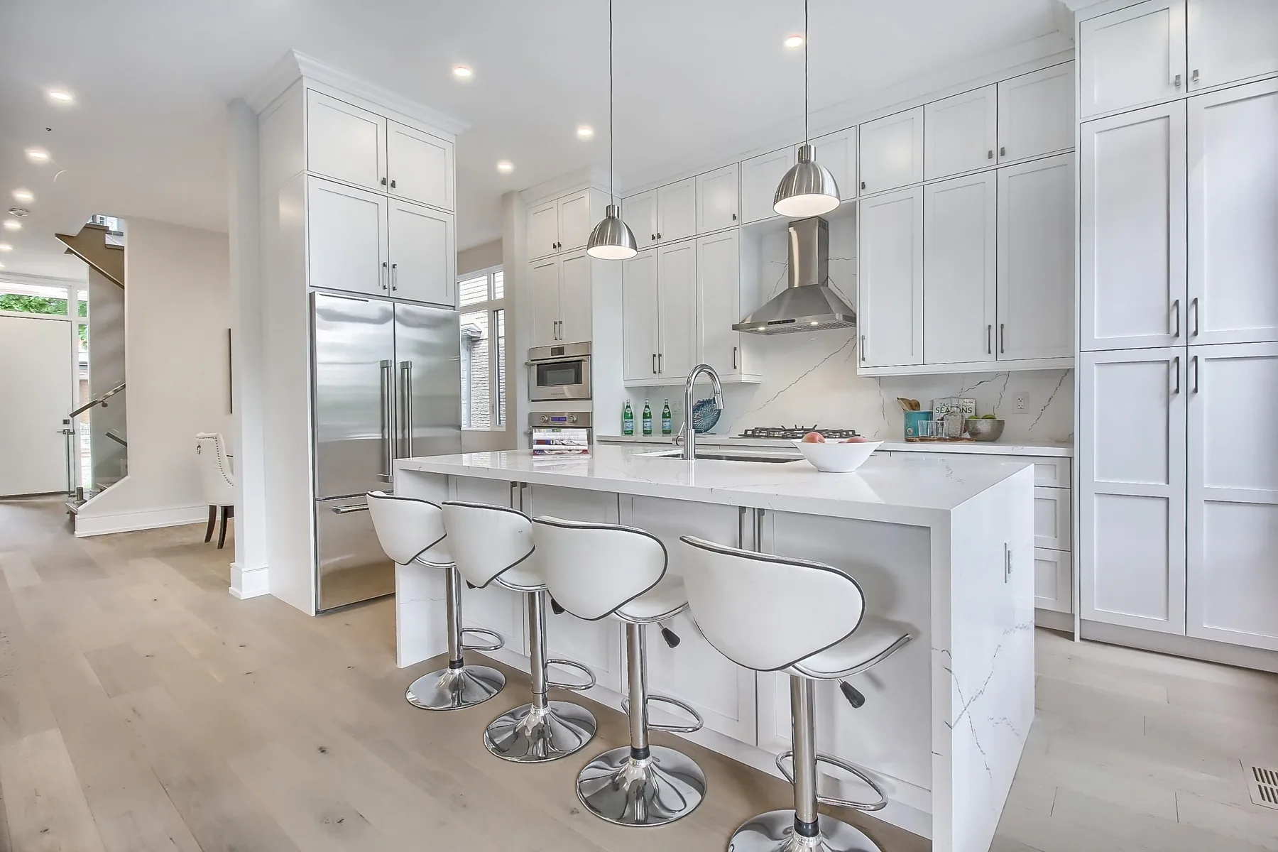 The kitchen from the far side, with barstools at the quartz island, full-height white cabinetry and a stainless range hood