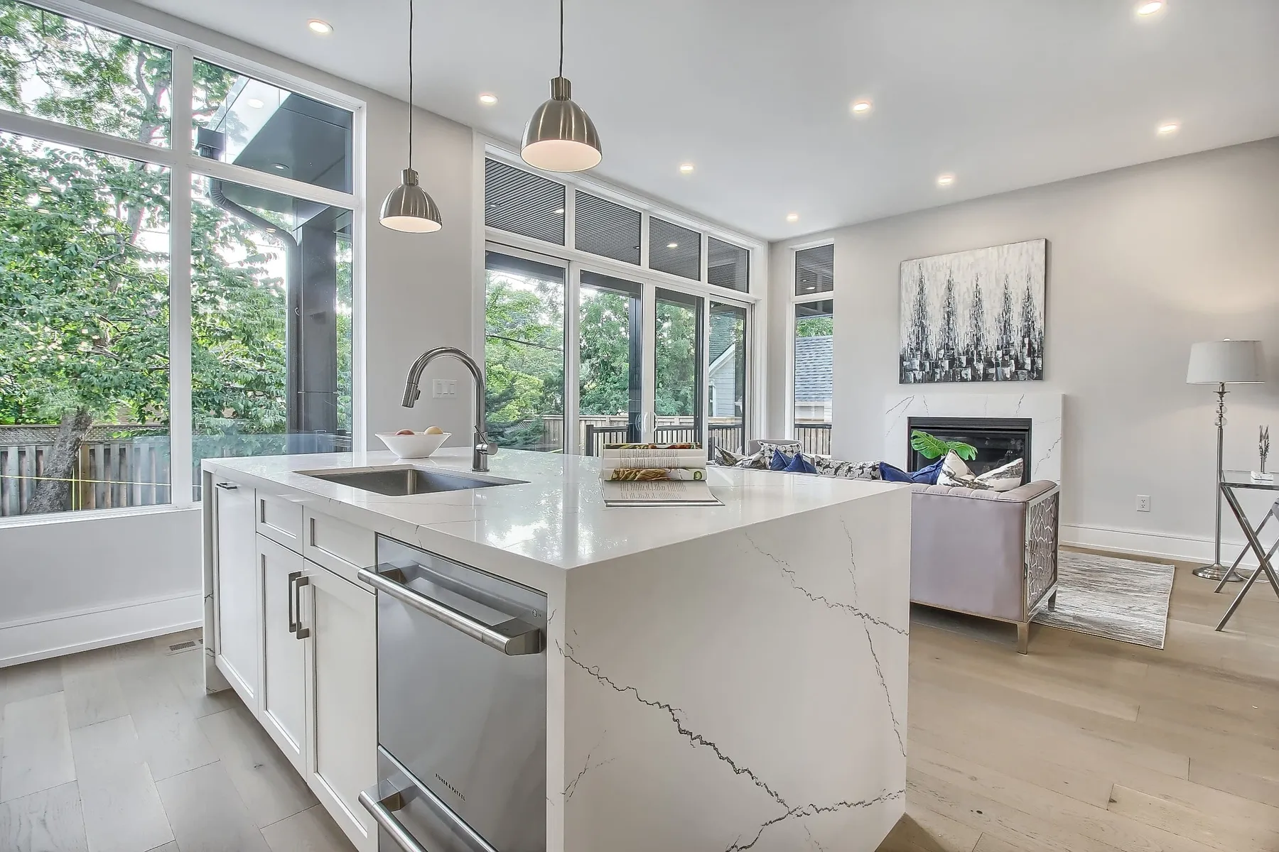 A white kitchen with a waterfall-edge quartz island, opening to a living room with a fireplace and floor-to-ceiling windows