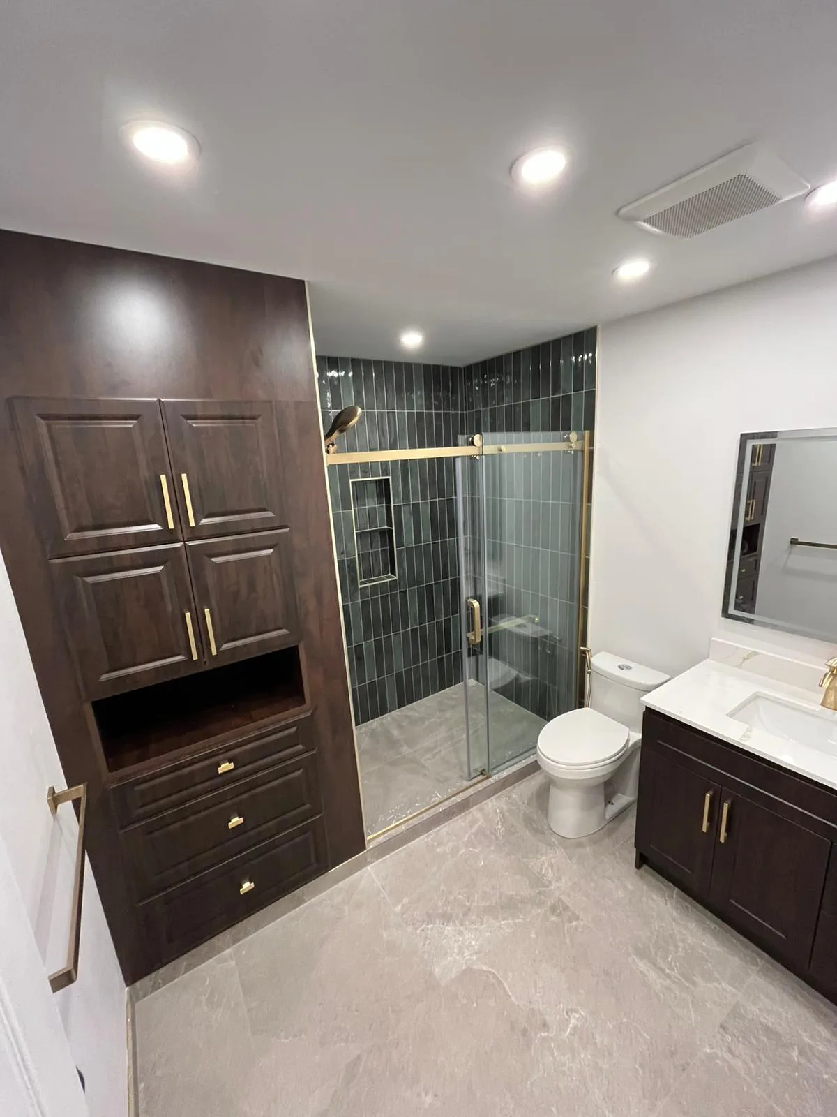 A renovated bathroom with a green-tiled glass walk-in shower, brass fixtures, walnut storage and a dark quartz-topped vanity